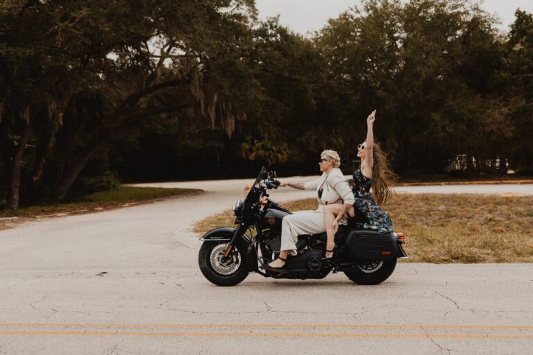 Two women ride a Harley-Davidson motorcycle, one steering while the other throws an arm up excitedly.