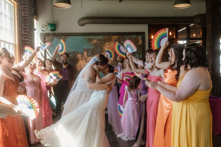 Two brides kiss while wedding party waves rainbow fans inside Tinker House, dressed in vibrant pink, orange, and yellow attire.