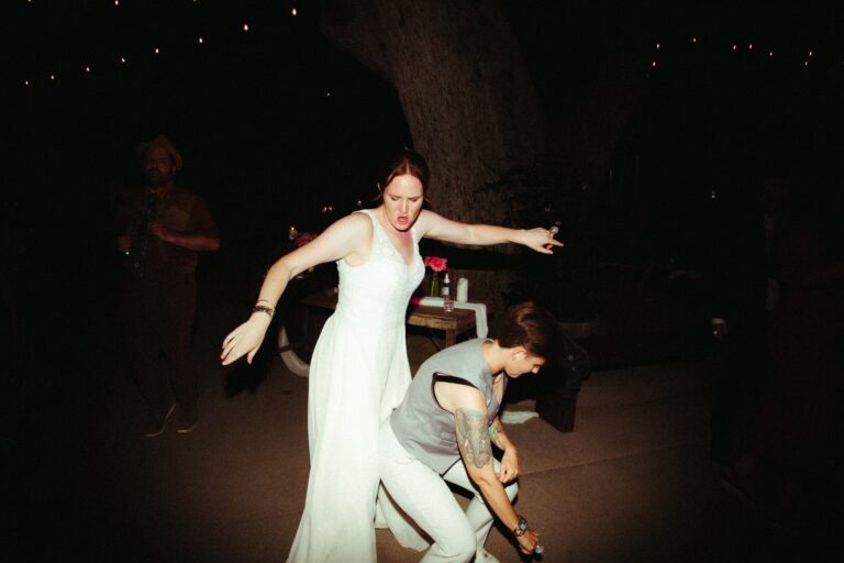 Bride in gown and bride in sleeveless vest dance playfully together at night reception.