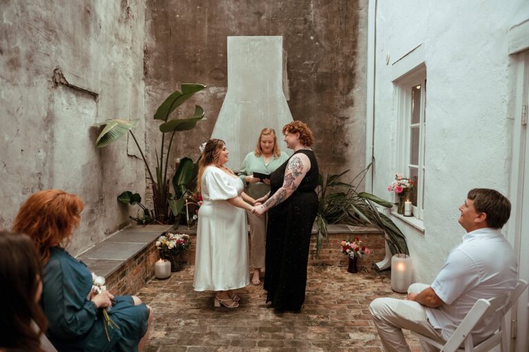 Jess and Cheyenne stand holding hands in a brick courtyard with officiant and seated guests.