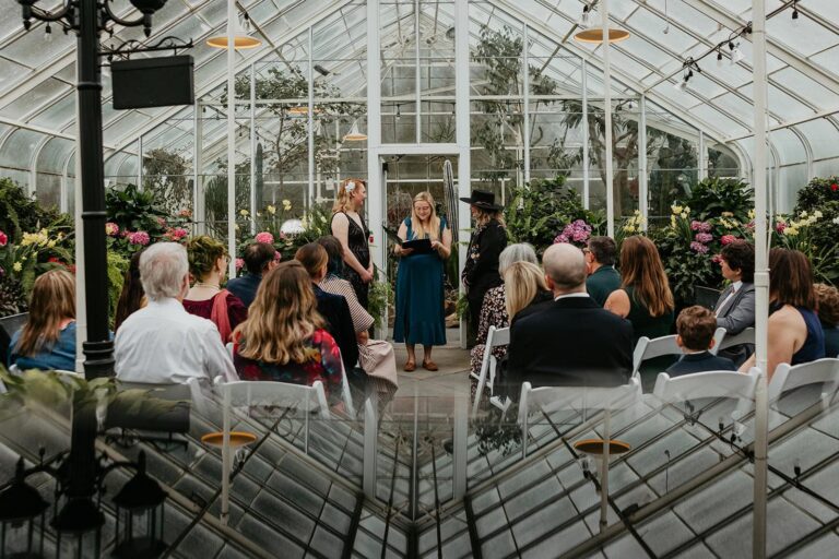 A small wedding ceremony takes place inside Volunteer Park Conservatory with guests seated and couple standing together.
