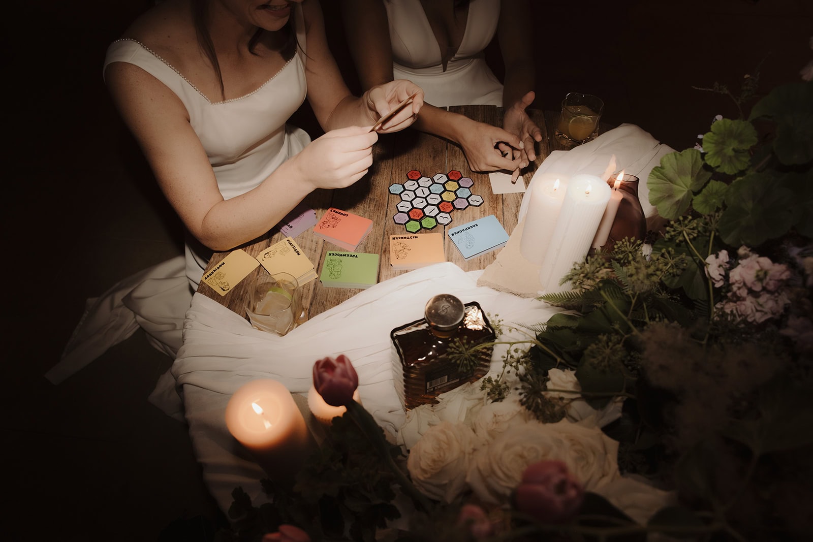 Two brides in white dresses sit at a wooden table playing a colorful card game by candlelight.