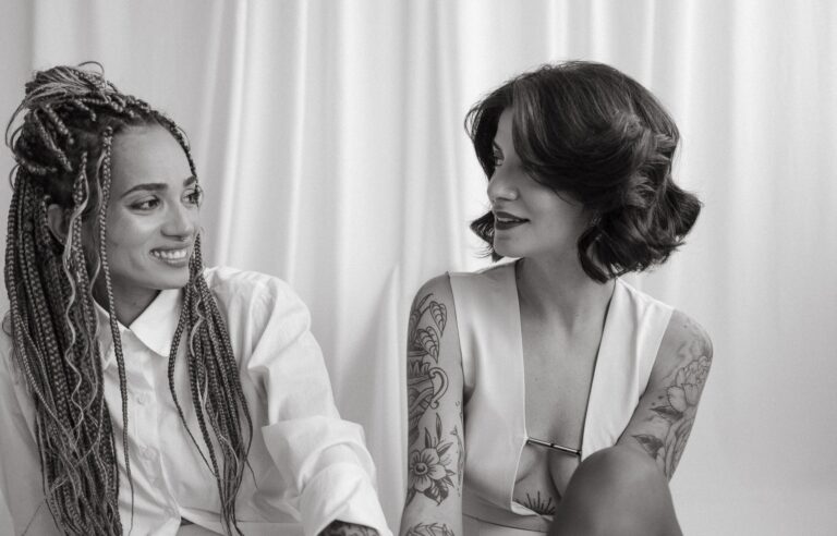 Two brides smile at each other, one with braids and shirt, the other with short dark hair and white dress.