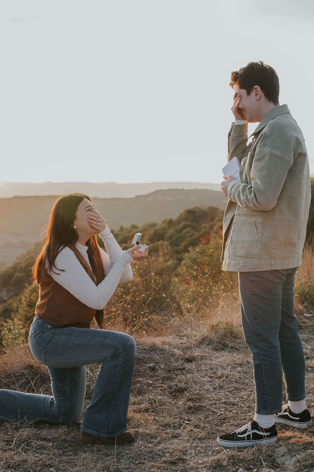 Woman kneels with ring box while her partner covers face in emotion during Catalonia proposal.