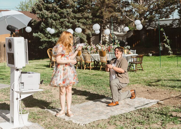 One partner kneels holding a ring box while the other holds photo booth strips at a backyard setup.