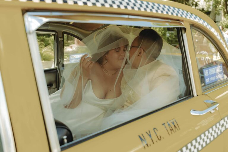 Two brides share a kiss under a veil while seated inside a vintage yellow NYC taxi.