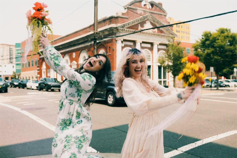 Two women in vintage 70s dresses laugh and hold floral bouquets on a street corner in Asbury Park.