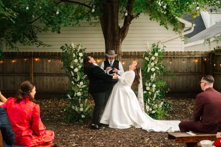 Two brides in gown and velvet jacket laugh together at altar surrounded by floral arches and string lights.
