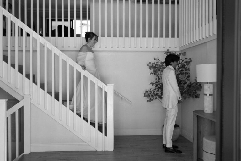 Bride in off shoulder gown walks down indoor staircase to meet her partner for their first look.