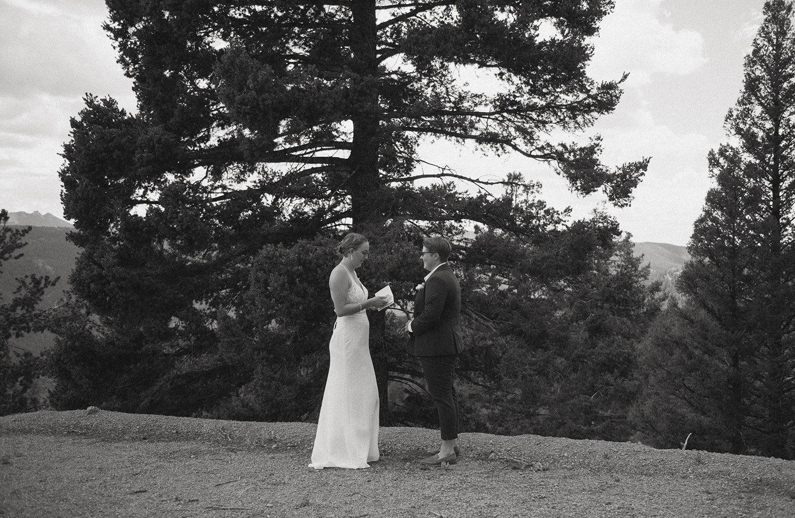 Two brides stand outdoors surrounded by tall pine trees as they exchange handwritten vows during their intimate ceremony.