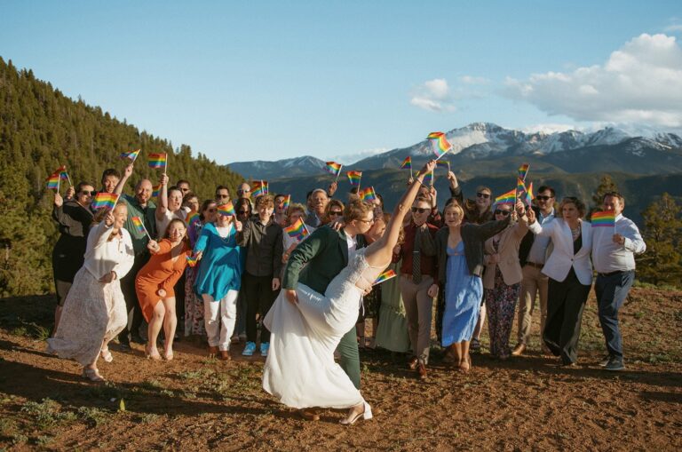 Two brides kiss while guests cheer and wave rainbow flags at their Colorado mountain wedding with Pikes Peak views.