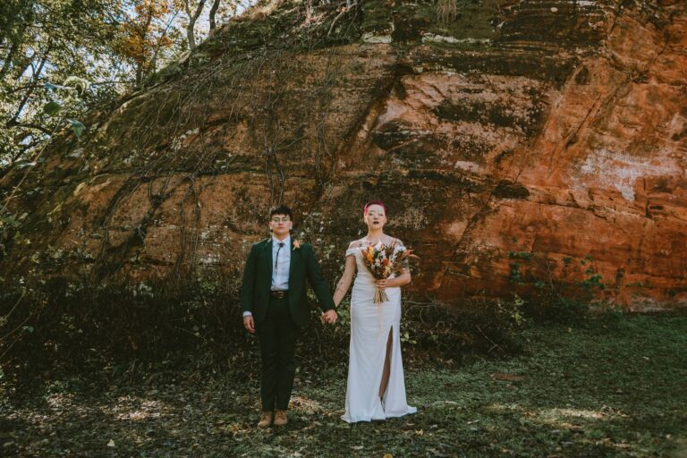 Couple holds hands in front of towering red rock wall, one wearing white gown, the other in green suit.