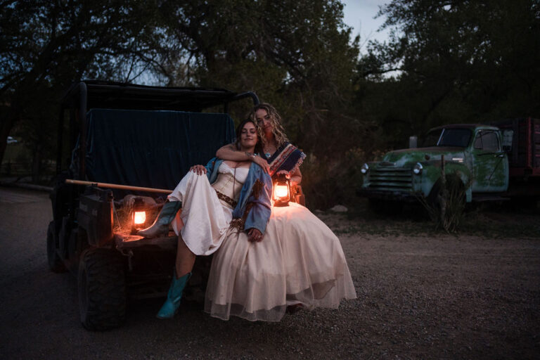 Two women sit together holding lanterns beside rustic ranch vehicle at night, vintage trucks in the background.