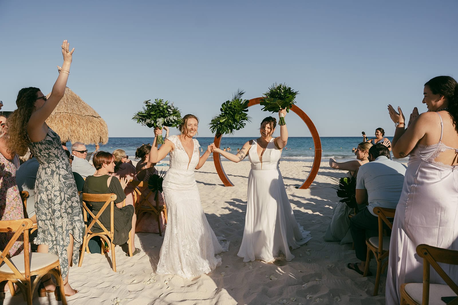 Two brides walk hand in hand down sandy aisle holding bouquets as guests cheer them on.