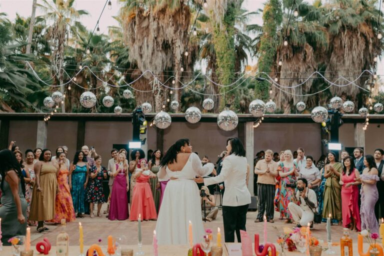 Couple shares first dance on outdoor dance floor under disco balls, surrounded by cheering wedding guests and string lights.