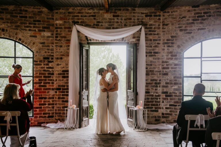 Two brides in satin gowns share their first kiss beneath rustic barn doors draped with flowing white fabric.