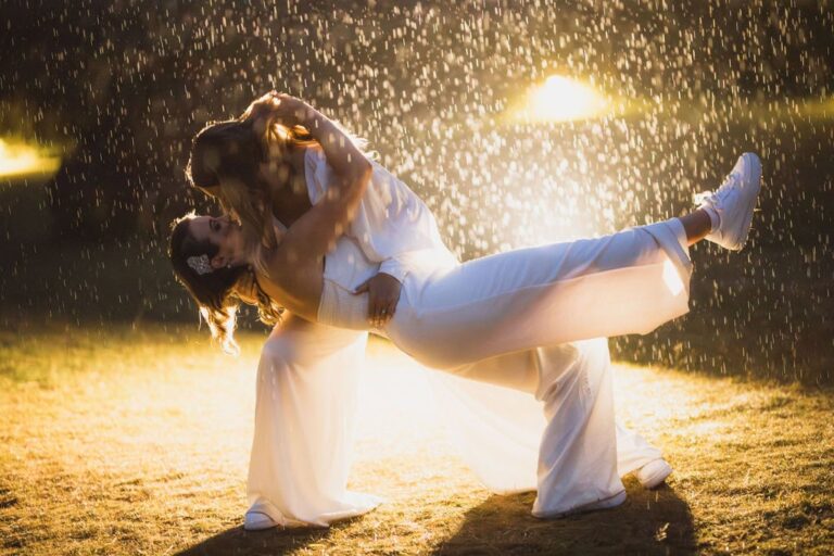 One bride dips the other in matching satin separates and sneakers, backlit by golden rain at night.