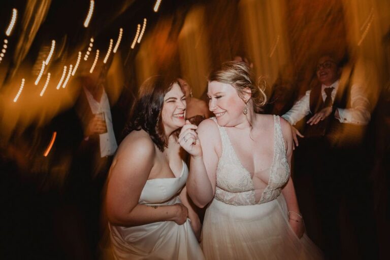 Two brides laugh and smile on dance floor surrounded by guests inside warmly lit wedding reception venue.