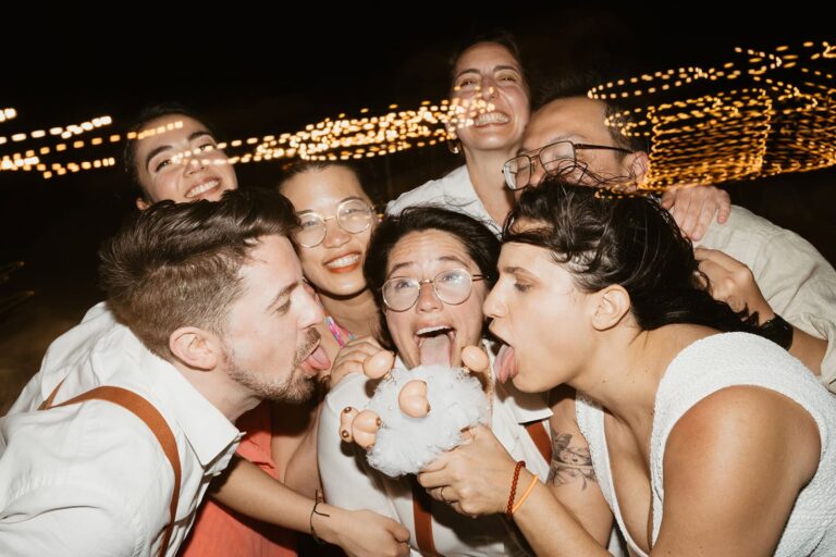 A group of wedding guests gather around the brides, laughing and playing with a novelty bouquet during the reception.
