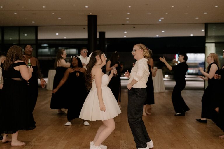Brides wearing white dress and dark trousers dance together in sneakers at indoor reception with guests.