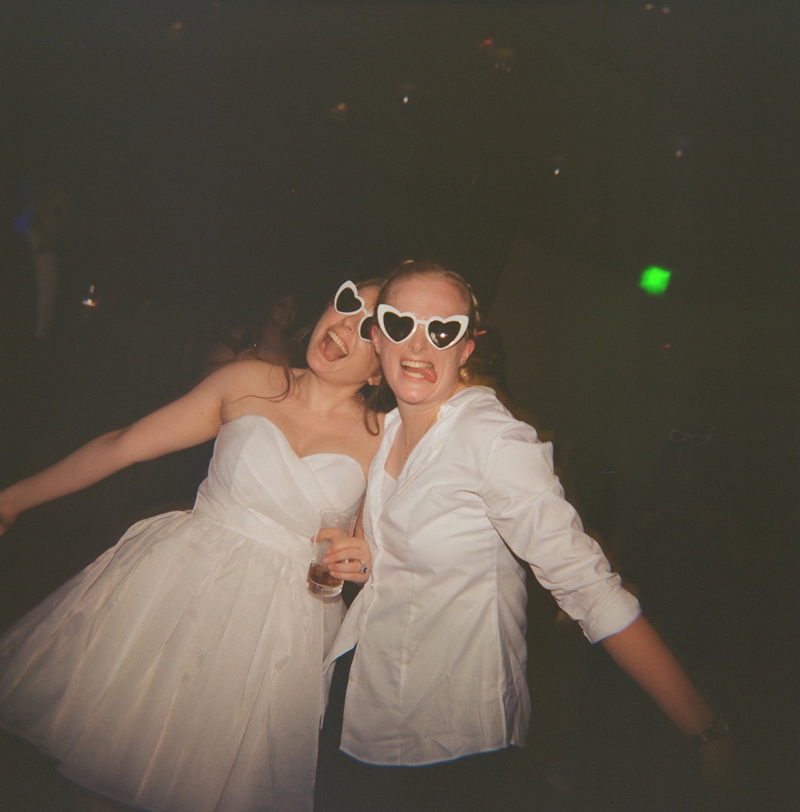 Bride in puff sleeve mini dress and partner in shirt and trousers pose together wearing white heart sunglasses.