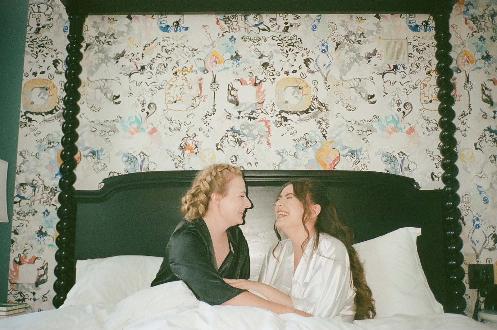 Brides wearing silk robes laugh together while sitting in patterned wallpaper hotel bed during wedding morning.