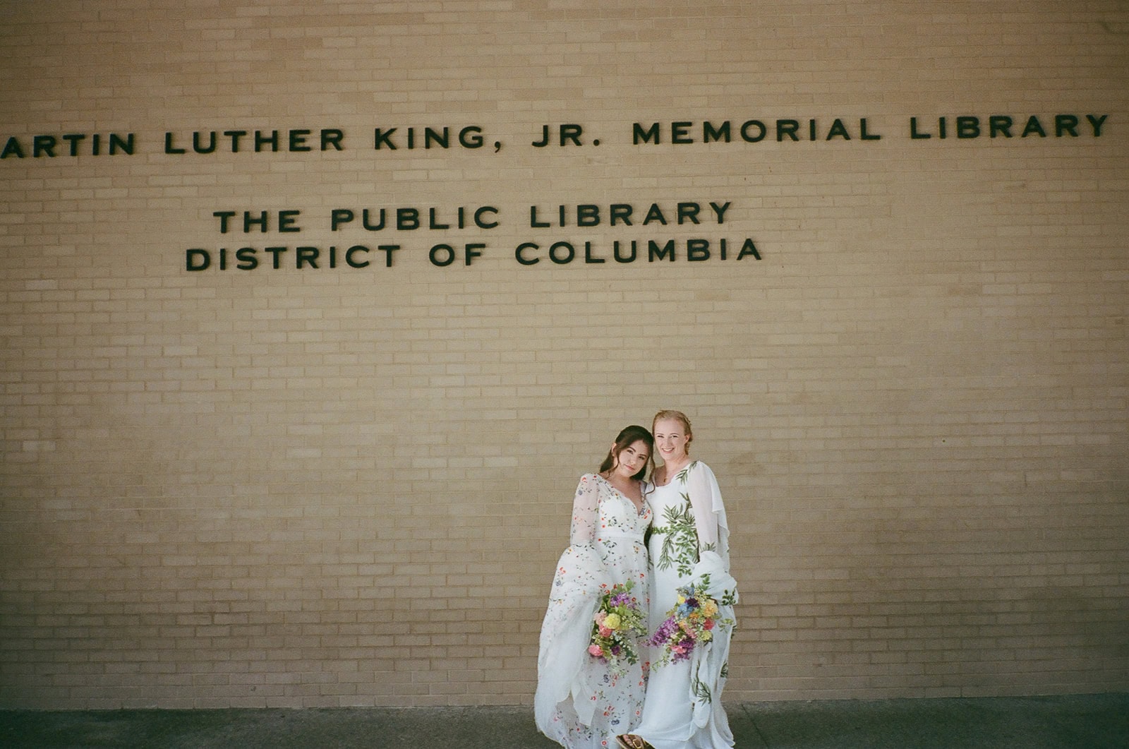 Brides in embroidered gowns stand together with bouquets against Martin Luther King Jr. Library exterior wall.