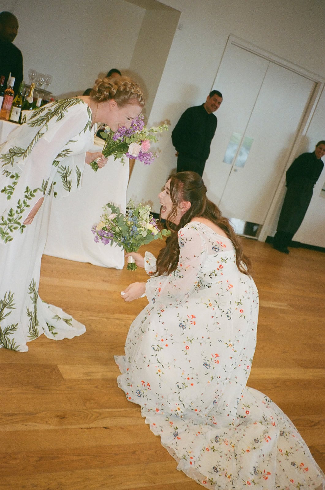 Bride in leafy embroidered gown laughs while holding bouquet as partner in floral gown kneels nearby.