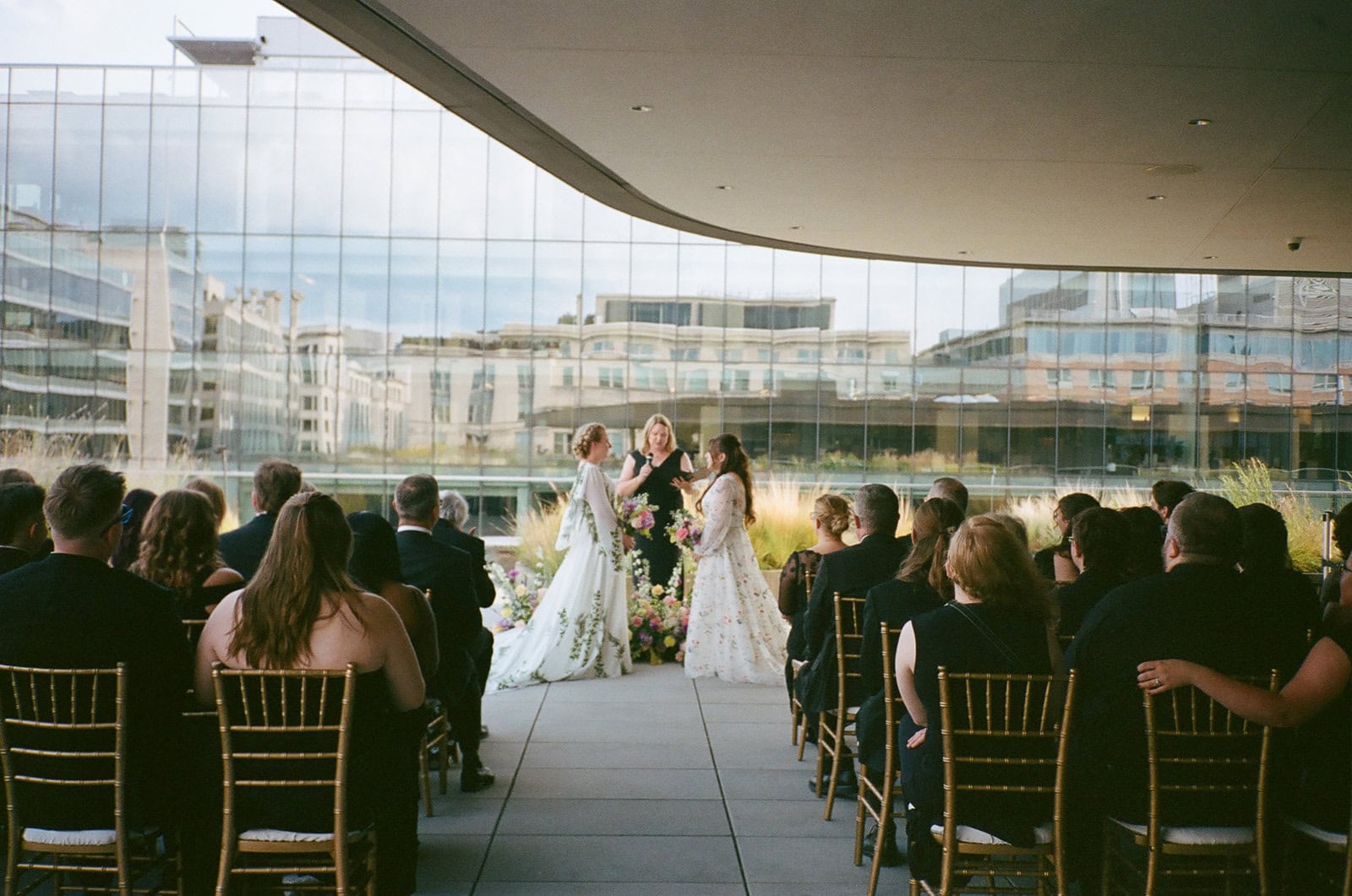 Brides in embroidered gowns stand with officiant during rooftop wedding ceremony facing seated guests.