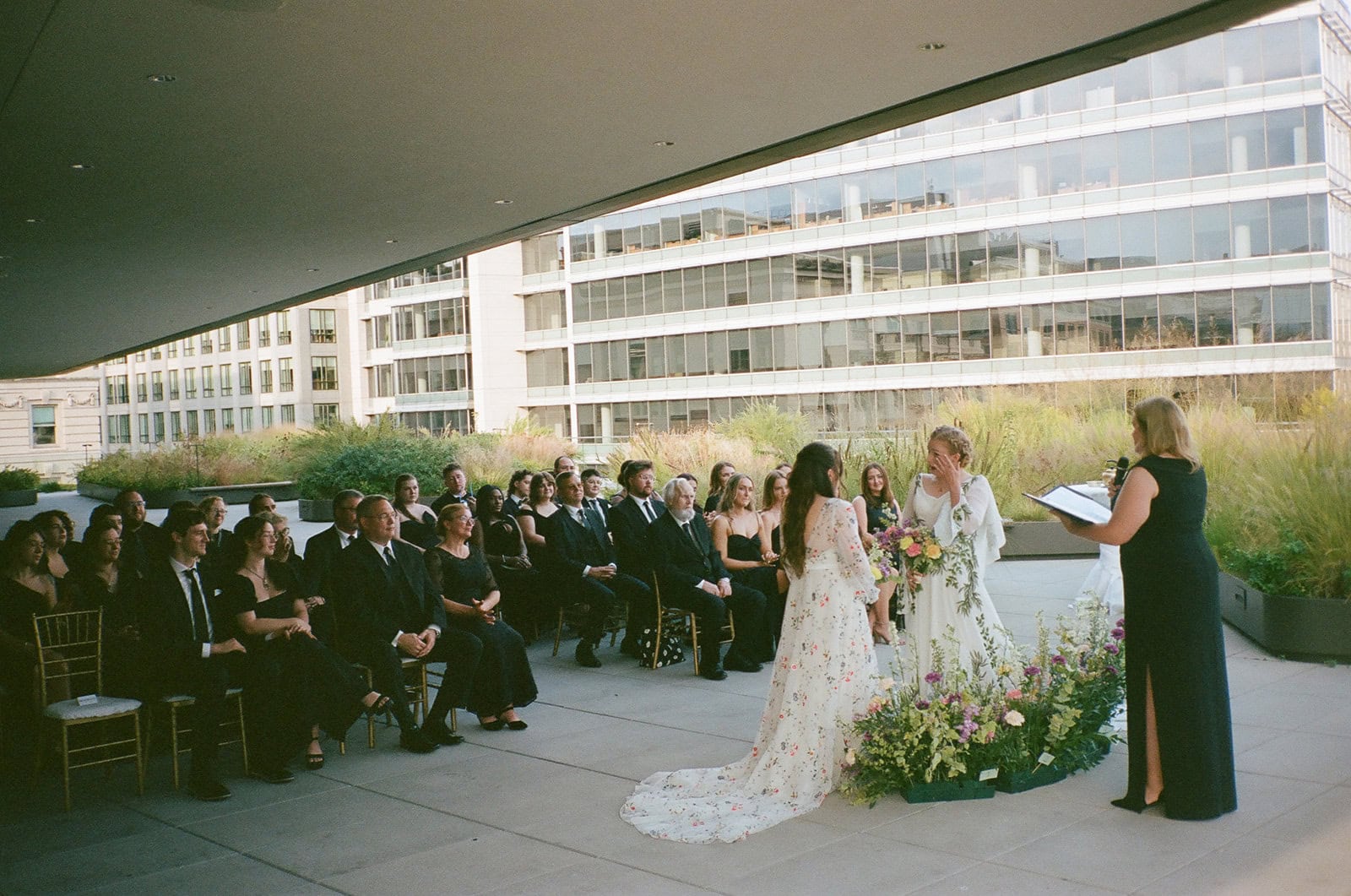 Bride in floral gown and bride in leafy gown exchange vows with officiant during rooftop ceremony.