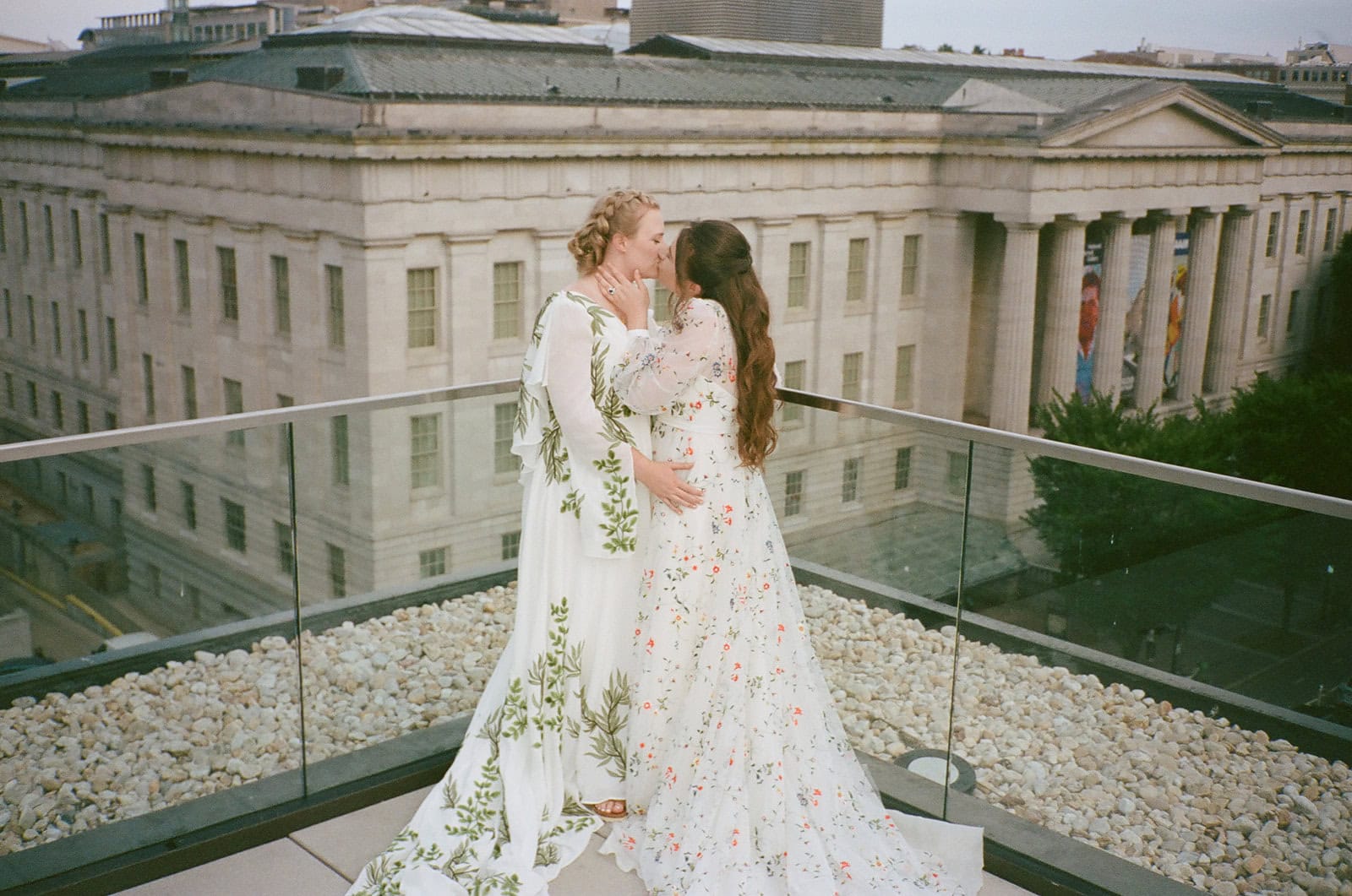 Brides in floral embroidered gowns kiss on rooftop terrace with Washington museum architecture in background.