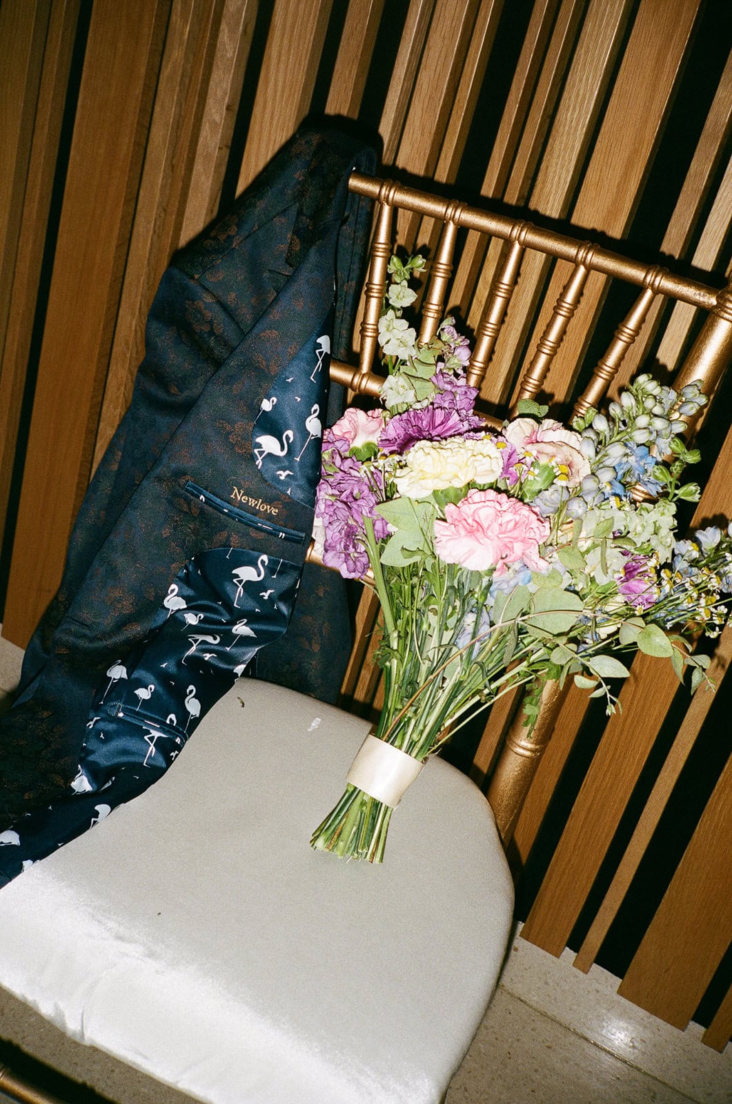 Colorful wedding bouquet rests beside custom suit jacket with flamingo lining on gold chair.