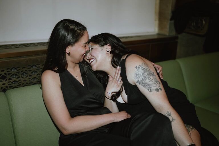 Two women in black outfits sit on green bench inside courthouse laughing closely together with bouquet nearby.