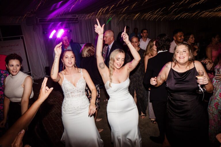 Two brides in gowns dance with guests on marquee dance floor with bright purple lighting.