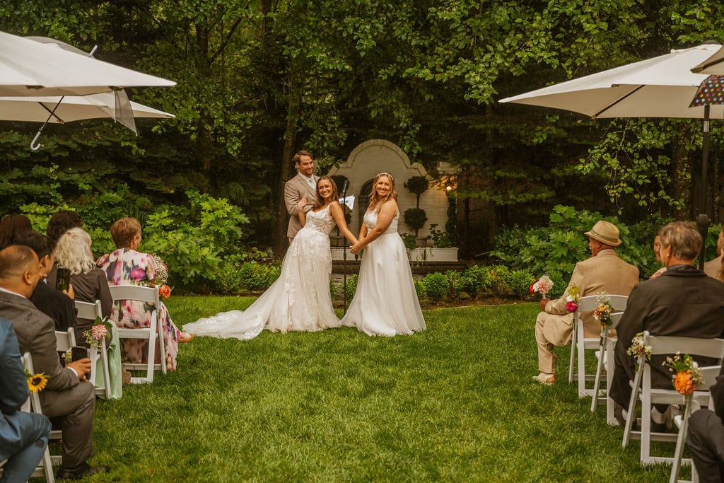 Two brides in lace gowns stand hand in hand with officiant outdoors surrounded by seated wedding guests.
