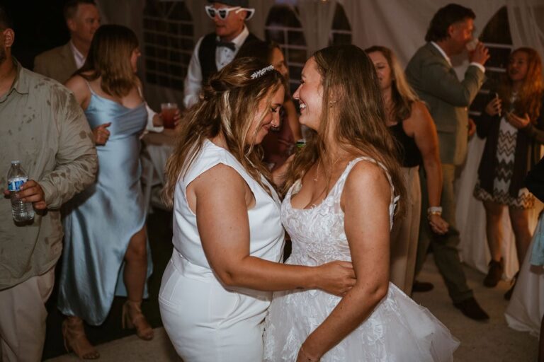 Two brides dance together on the reception floor surrounded by guests, one wearing a lace gown and one in white suit.