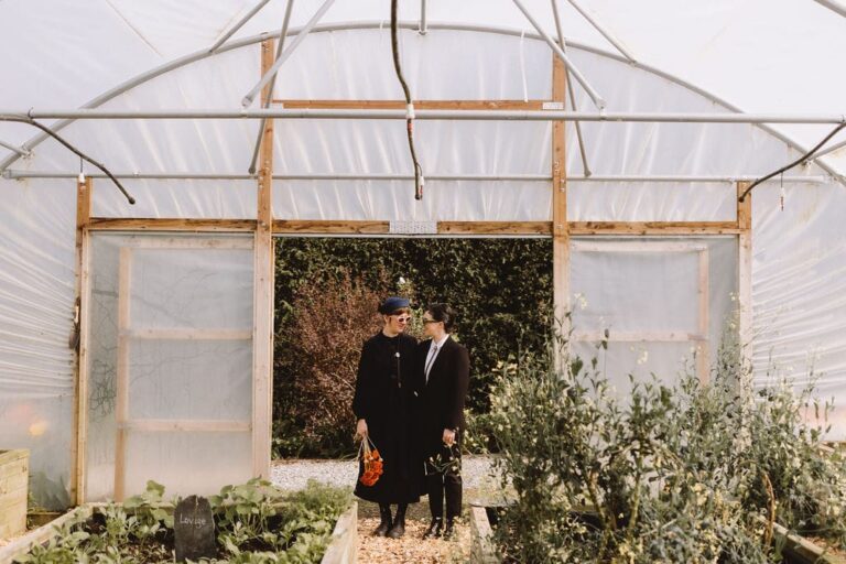 Brides in dark outfits walk through greenhouse polytunnel surrounded by garden plants and raised beds.