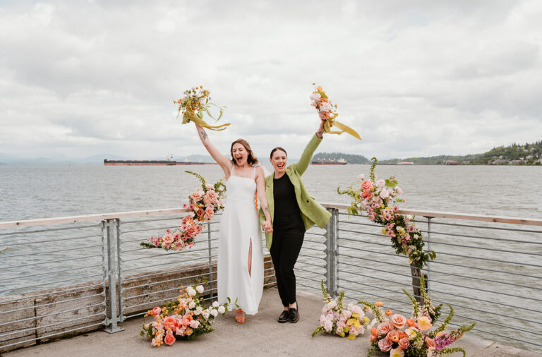 A queer couple celebrates at their waterfront ceremony, raising colorful bouquets above their heads with happiness.