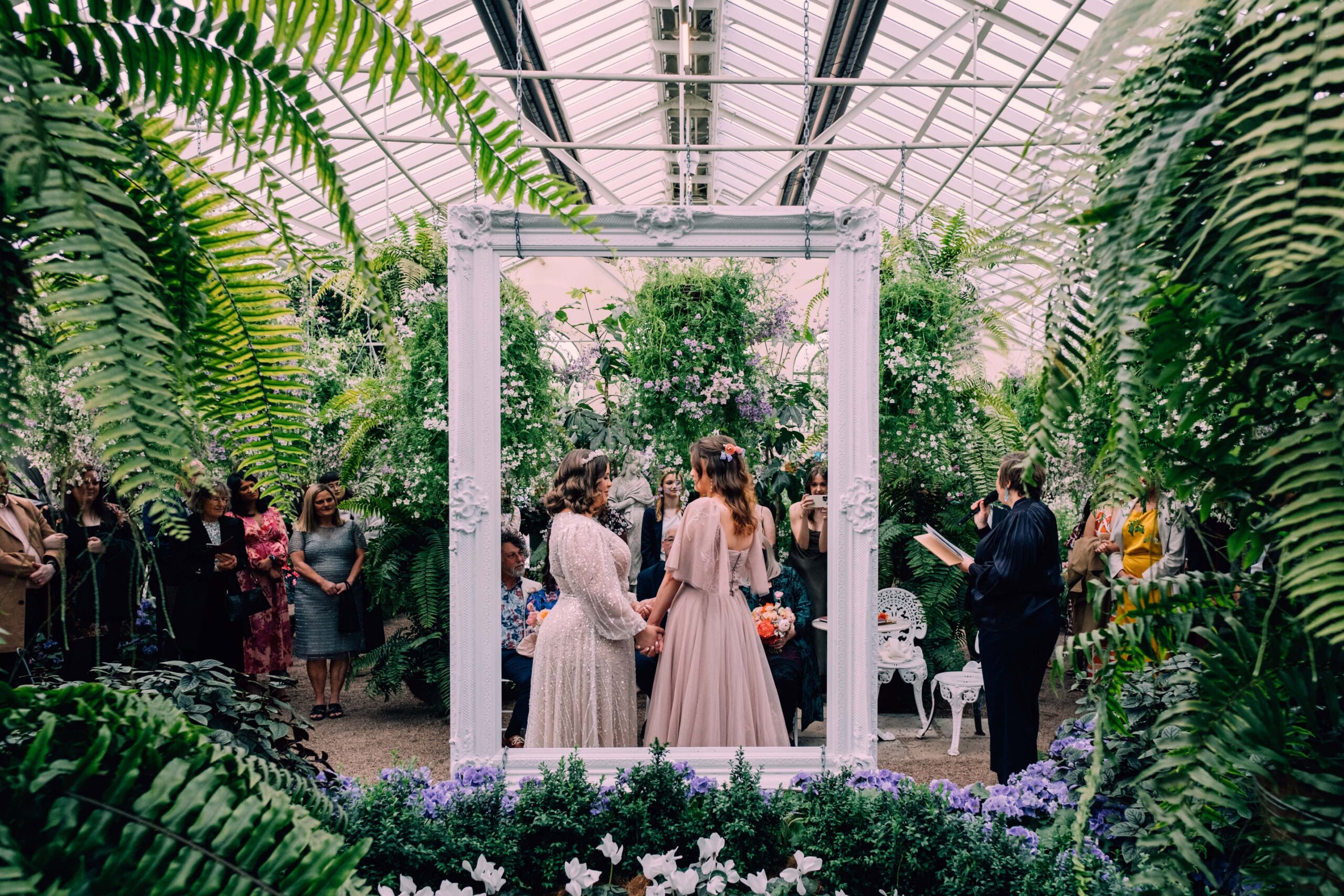 Two brides stand holding hands beneath hanging greenery during their ceremony framed by a white arch.