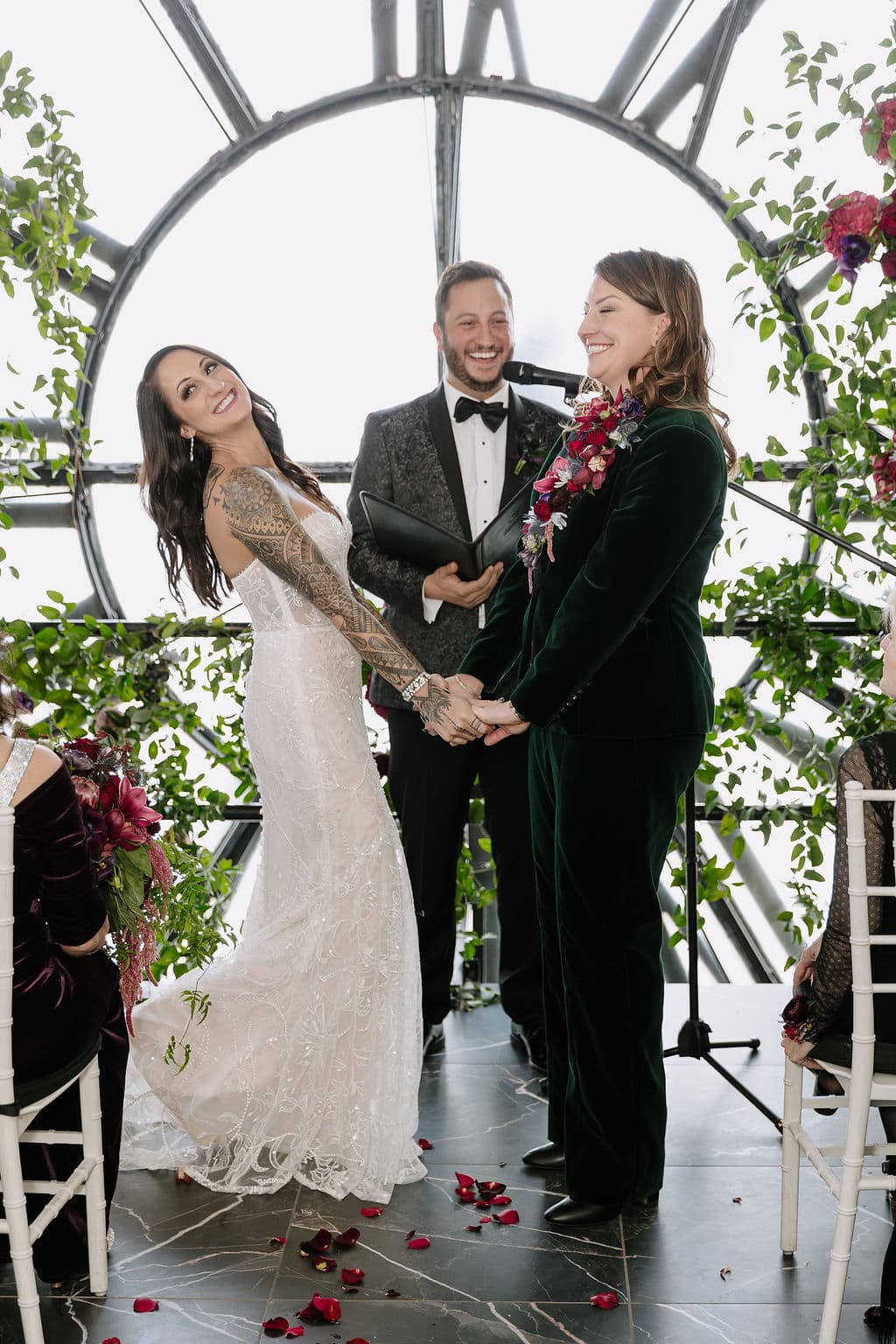 A bride in a lace gown and her partner in a velvet suit hold hands during vows.