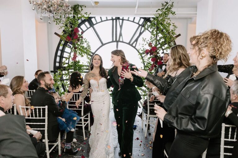 Two brides walk down the aisle together as guests cheer and throw flower petals in celebration.