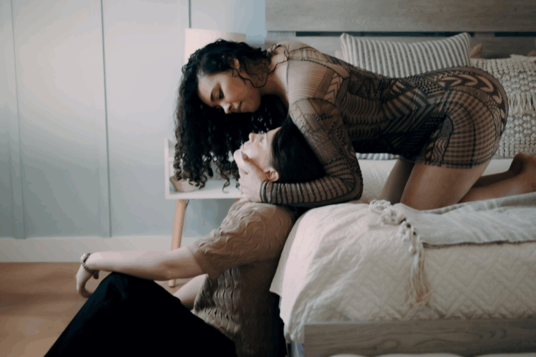 One woman kneels on a bed while leaning down toward her partner, who sits on the floor beside her.