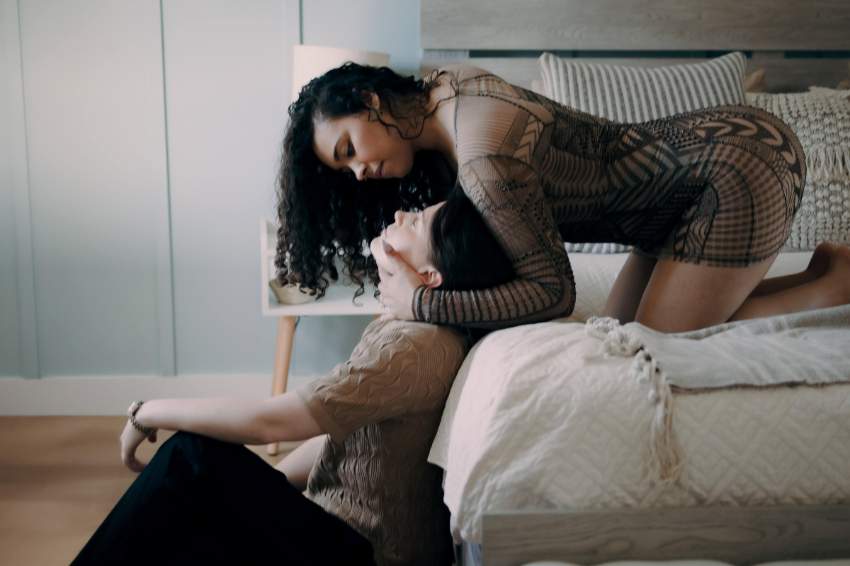 One woman kneels on a bed while leaning down toward her partner, who sits on the floor beside her.
