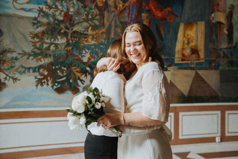 A couple hug and laugh in front of a colorful mural, one holding a bouquet of white roses