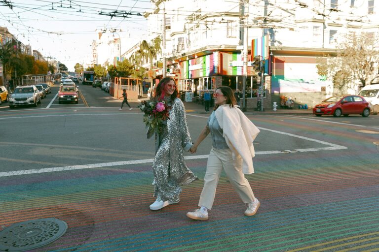 Two brides holding hands cross the Castro’s rainbow-striped crosswalk with colorful storefronts and city traffic behind them.