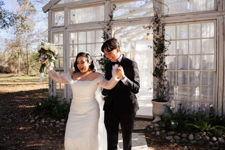 Newly married brides walk out of glasshouse hand in hand, smiling and raising bouquet under sunlight.