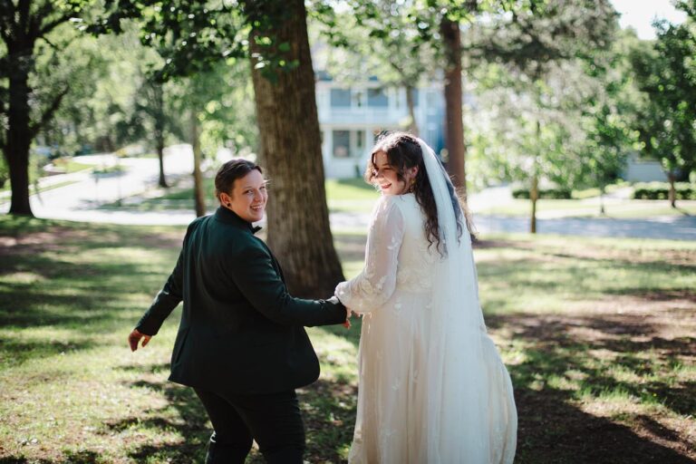 Two newlyweds walk hand in hand through a park, smiling back toward the camera under leafy trees.