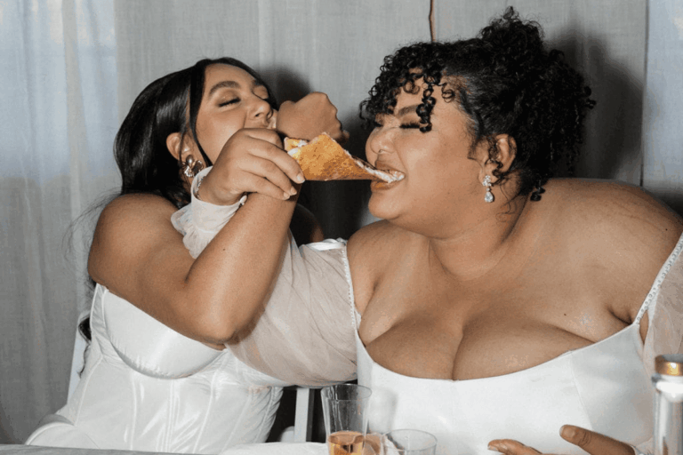 A couple in white laugh as one feeds the other a slice of pizza at their DIY Indiana wedding