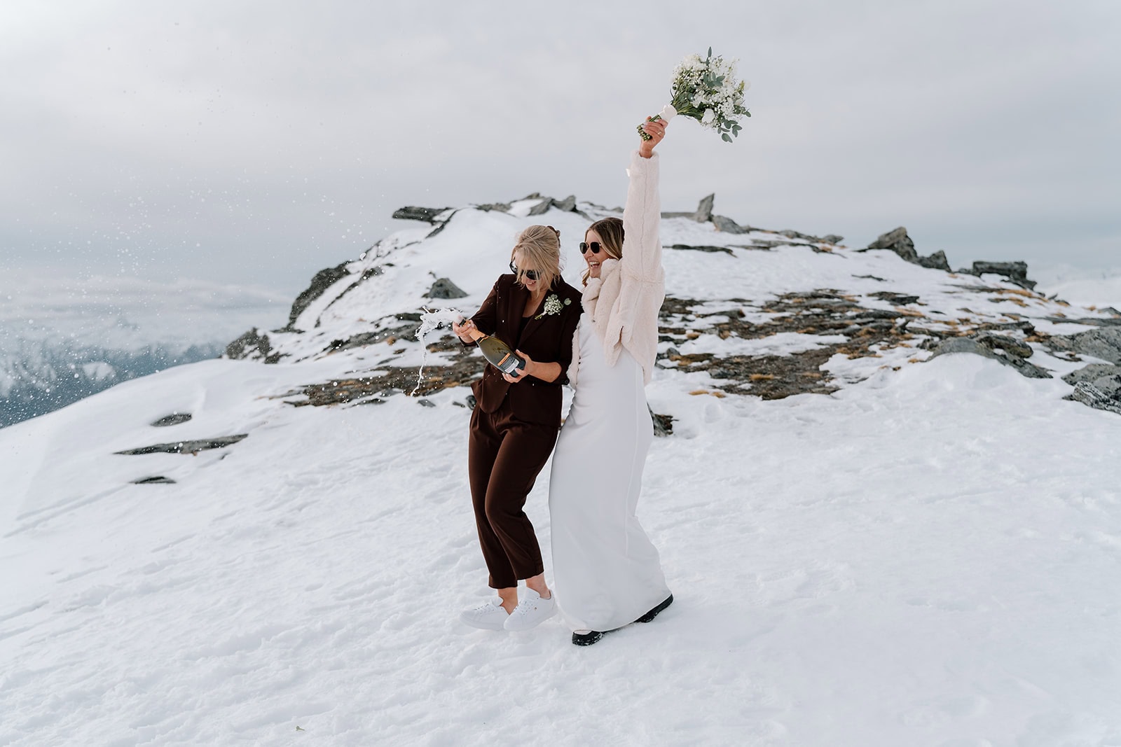 A couple celebrate on a snowy mountain peak in Queenstown's Remarkables, one raising a bouquet in the air