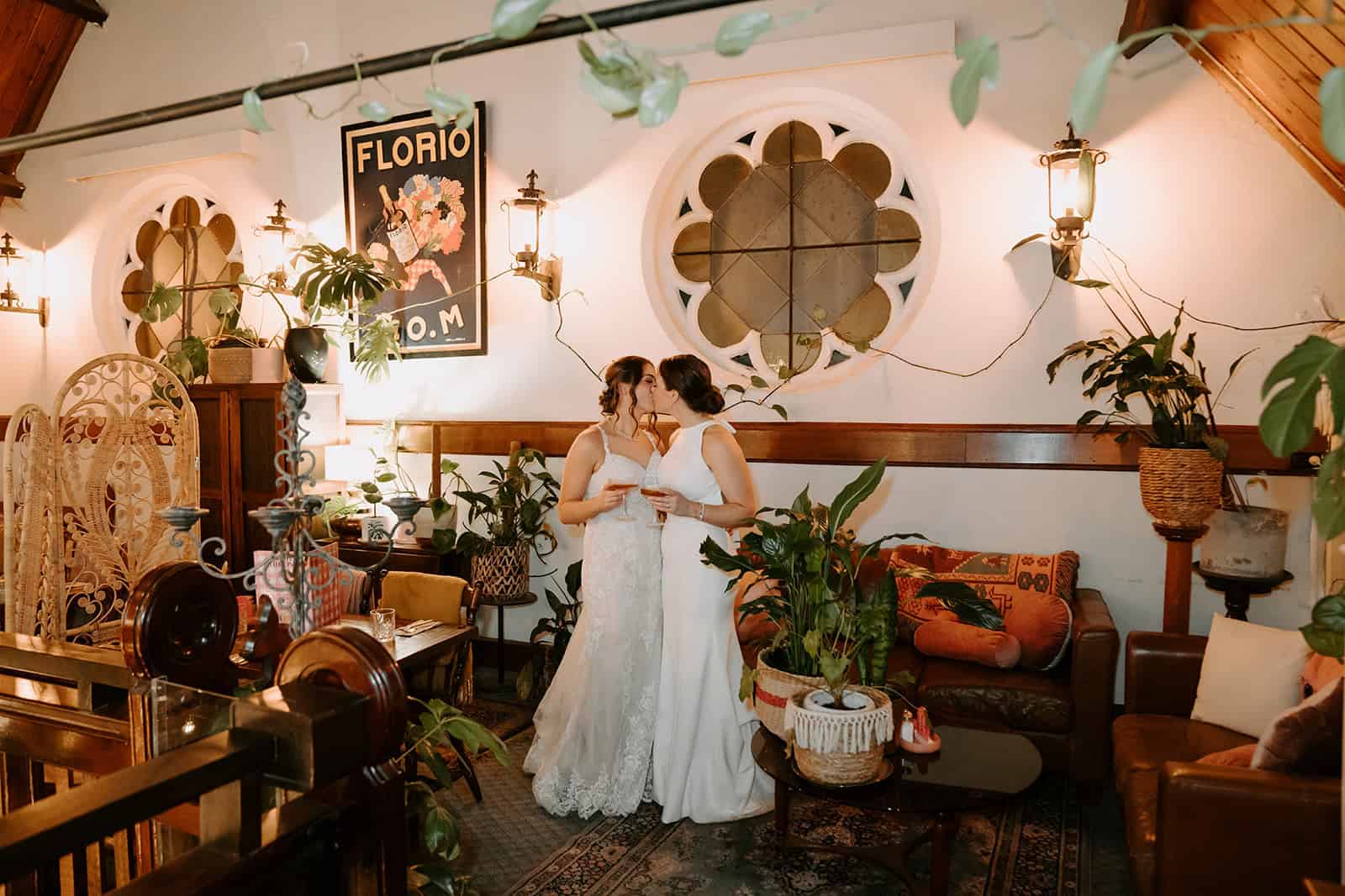 A couple in white stand together in a plant-filled venue with vintage furniture and a round window at their Lilydale wedding